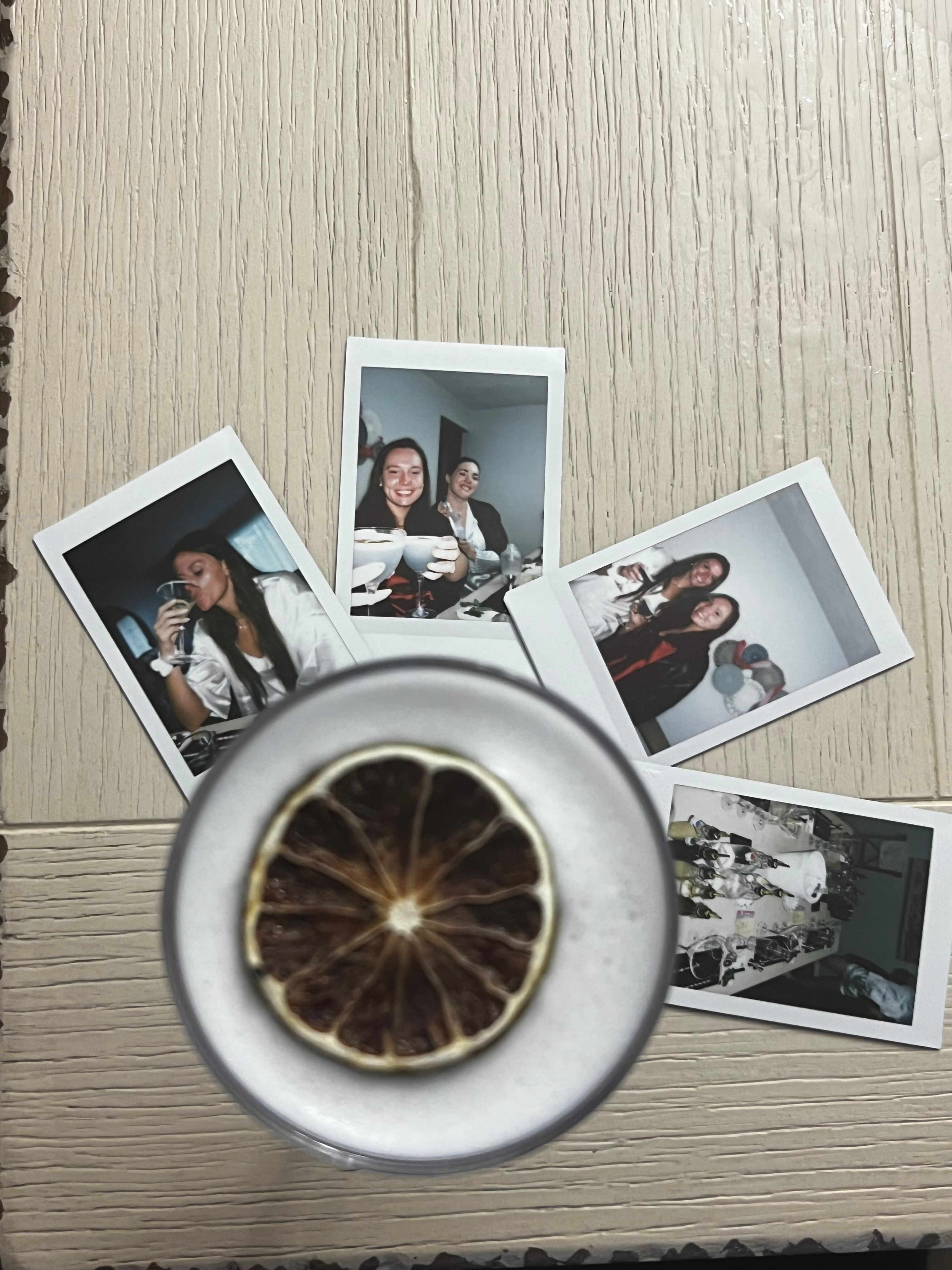 Flatlay of five Polaroid instant photos spread on a light wood table with a frothy cocktail topped by a dried lime slice in the foreground — party snapshots and drinks.