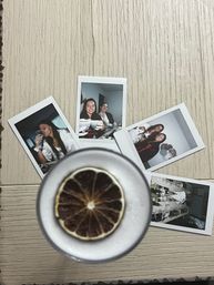 Flatlay of five Polaroid instant photos spread on a light wood table with a frothy cocktail topped by a dried lime slice in the foreground — party snapshots and drinks.