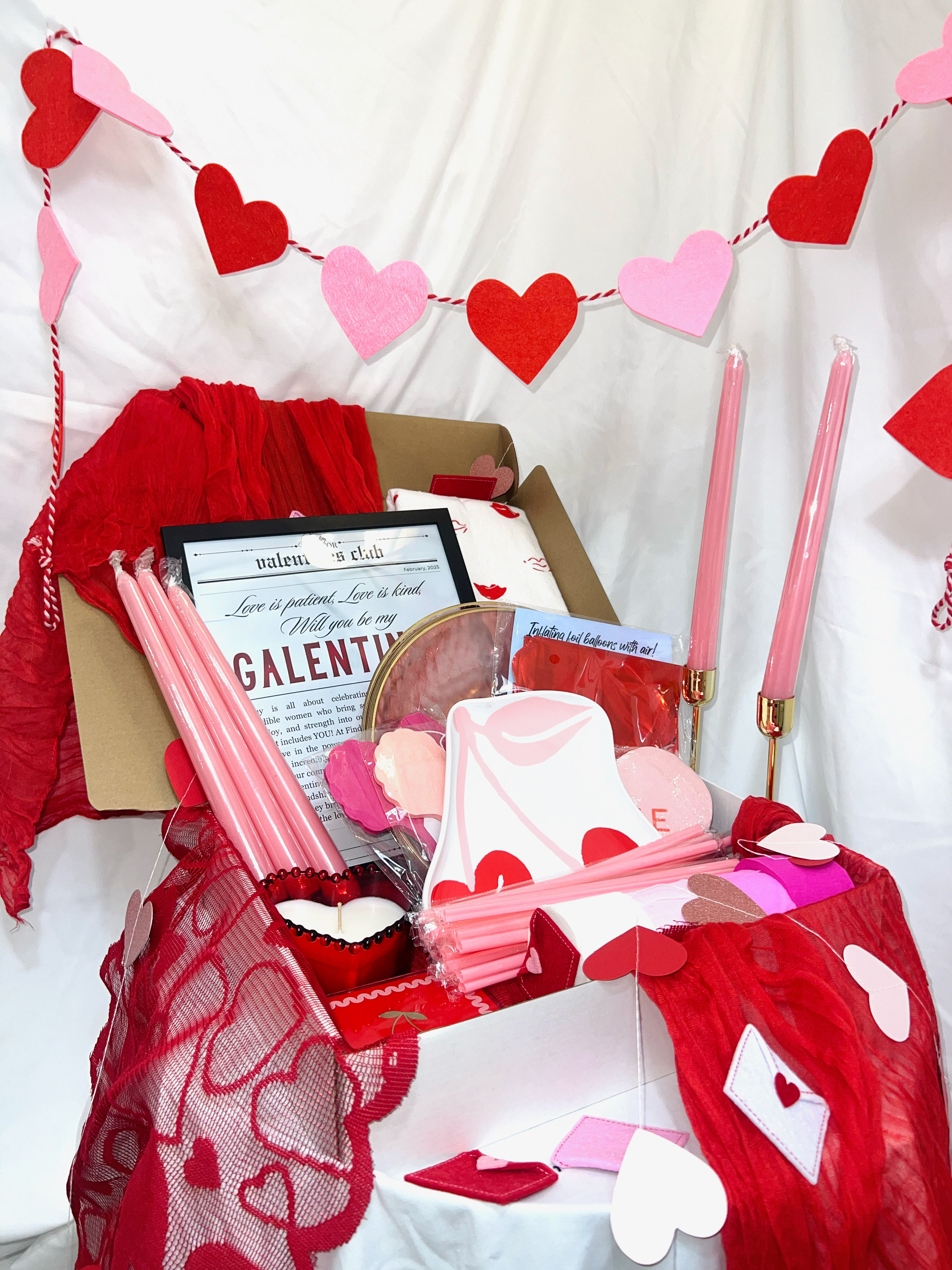 Galentines-themed Valentine gift box with pink taper candles, red tissue, heart garland, scattered heart confetti and paper treats on a white backdrop