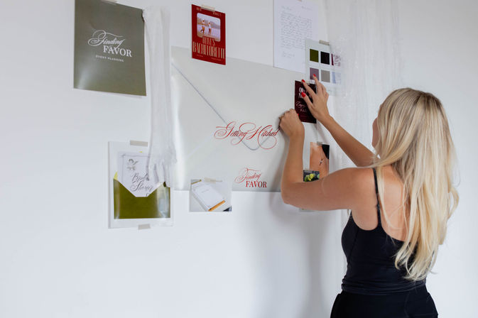 Person with long blonde hair pinning a wedding inspiration mood board with invitations, color swatches and photos on a white wall in a home studio.