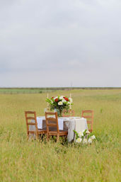 Rustic wooden table with four chairs, gingham tablecloth, brass candlesticks and a colorful floral centerpiece set in a grassy countryside meadow under a cloudy sky