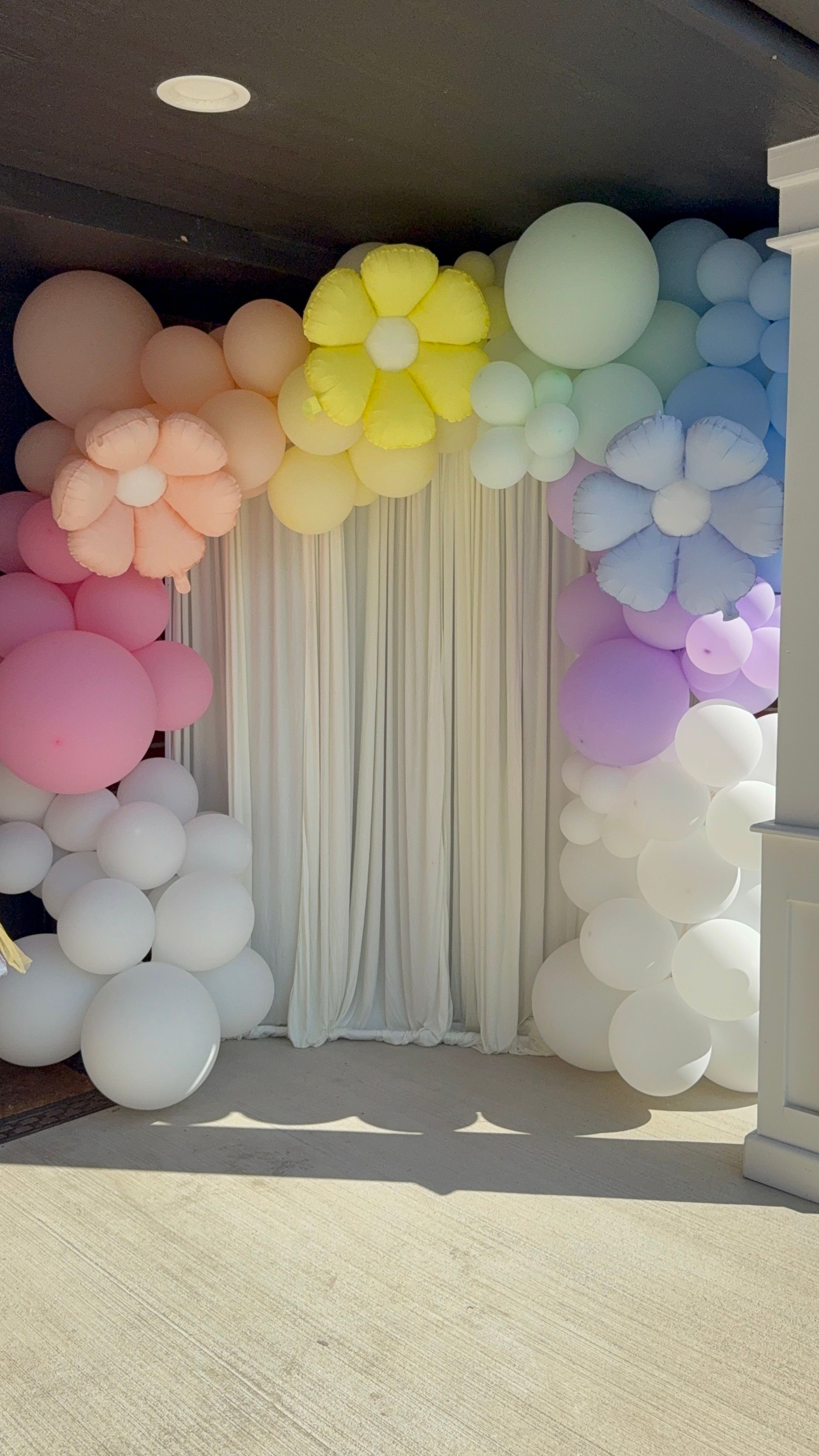 Pastel rainbow balloon arch with oversized flower-shaped balloons framing a white draped curtain photo backdrop at an outdoor event entrance