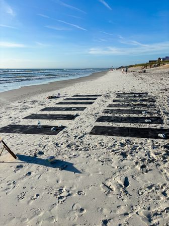 Rows of black yoga mats laid out on a sunny oceanfront sandy beach with gentle waves and a bright blue sky, people strolling in the distance — a beach yoga class setup.