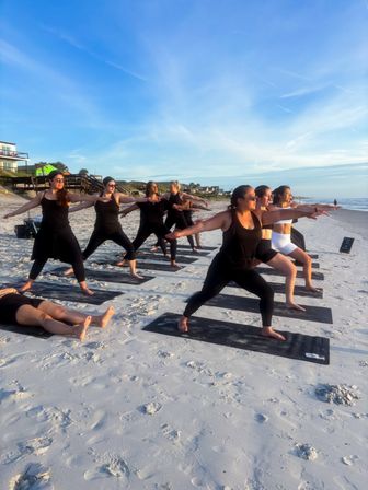 Sunlit beach yoga class on a sandy oceanfront shore — a group practicing warrior poses on mats near houses and a boardwalk at golden hour.