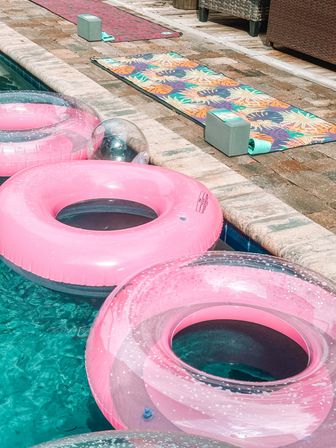 Backyard poolside scene with pink inflatable swim rings floating in turquoise water alongside a stone paver deck with colorful tropical-patterned mats and foam blocks
