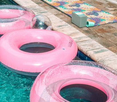 Backyard poolside scene with pink inflatable swim rings floating in turquoise water alongside a stone paver deck with colorful tropical-patterned mats and foam blocks