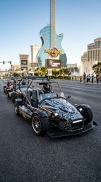 Line of black open-frame go-karts parked on the Las Vegas Strip with a giant guitar-shaped hotel tower and fountain in the sunlit background