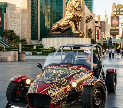 Eye-catching open-top sports kart with red-and-gold graphics parked on the Las Vegas Strip, golden lion statue and green high-rise hotel in the background with pedestrians strolling.