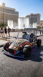Brightly painted open-frame go-kart with casino-themed graphics parked on the Las Vegas Strip in front of large dancing hotel fountains and resort towers on a sunny day.