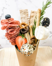 Gourmet charcuterie cone bursting with folded salami rosettes (blueberry pick), mini brie topped with a blackberry, everything-seasoned cheese ball, halved strawberry, pretzels (one chocolate-covered), breadsticks, crackers, walnuts and a rosemary sprig in a kraft paper cup.
