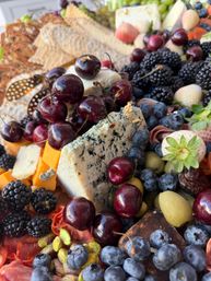 Vibrant gourmet charcuterie board close-up: blue cheese wedge topped with cherries, surrounded by blueberries, blackberries, cheddar cubes, olives, pistachios, crackers and cured meats.