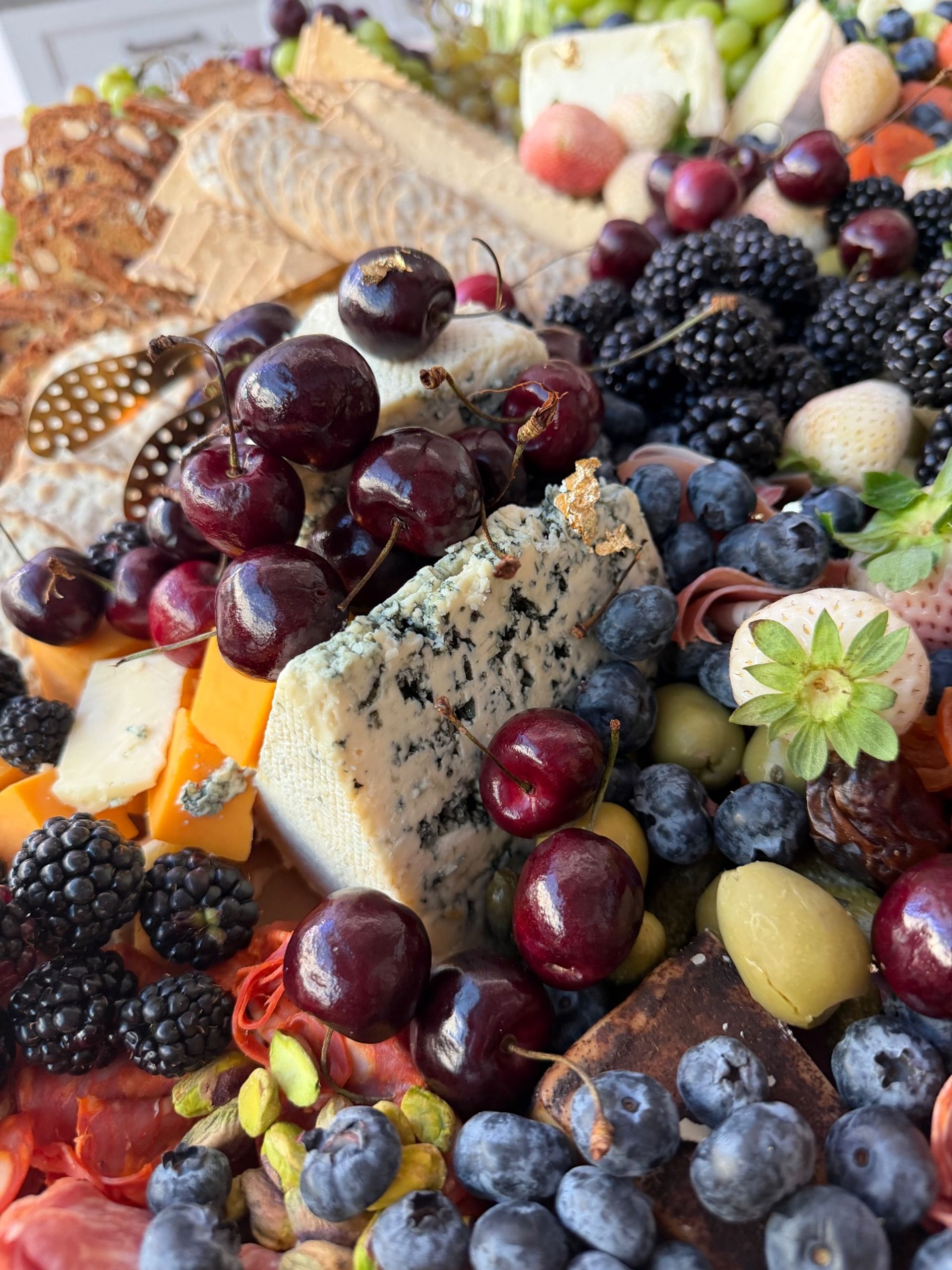 Vibrant gourmet charcuterie board close-up: blue cheese wedge topped with cherries, surrounded by blueberries, blackberries, cheddar cubes, olives, pistachios, crackers and cured meats.
