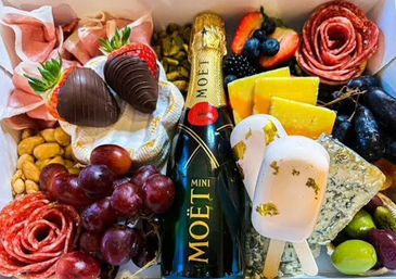 Gourmet charcuterie gift box with a mini Moët champagne bottle surrounded by assorted cheeses (blue, cheddar), salami roses, chocolate-covered strawberries, red grapes, olives, nuts and gold-leaf dessert popsicles — luxe party platter.