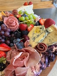 Close-up of a vibrant charcuterie board on wood: prosciutto and salami rosettes, blue cheese with edible gold leaf, a creamy cheese wedge, green and red grapes, strawberries, blueberries, blackberries and cherries.