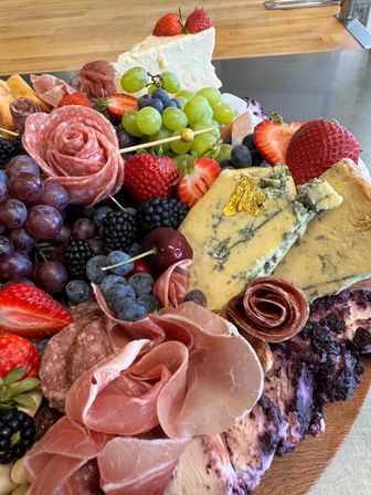 Close-up of a vibrant charcuterie board on wood: prosciutto and salami rosettes, blue cheese with edible gold leaf, a creamy cheese wedge, green and red grapes, strawberries, blueberries, blackberries and cherries.