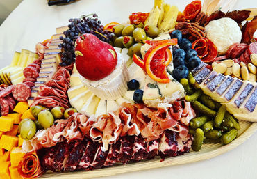 Gourmet charcuterie board with a brie wheel topped by a red pear, blue cheese and cheddar slices, rolled prosciutto and salami, green olives, cornichons, pecans and almonds, grapes, blueberries and blood orange slices.
