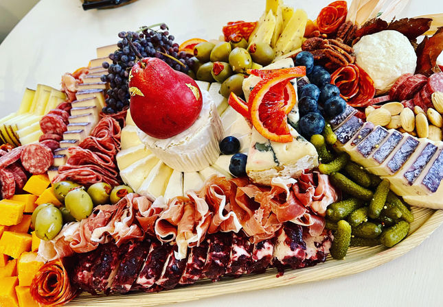 Colorful gourmet charcuterie board on a wooden platter with assorted cheeses (brie, blue, aged slices), cured meats (prosciutto, salami), green olives, cornichons, grapes, blueberries, a red pear, blood orange slices and nuts.