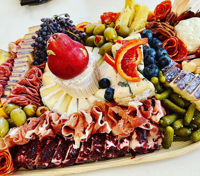 Gourmet charcuterie board with a brie wheel topped by a red pear, blue cheese and cheddar slices, rolled prosciutto and salami, green olives, cornichons, pecans and almonds, grapes, blueberries and blood orange slices.