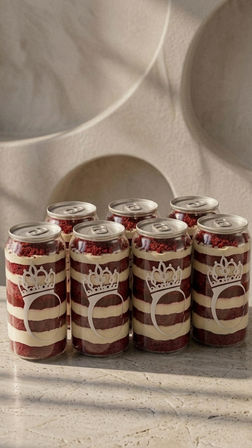 Six clear cans of layered red velvet cake and cream — cake-in-a-can desserts with pull-tab lids and crown motif — arranged on a marble countertop against a neutral textured wall.