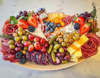 Vibrant gourmet charcuterie board on a wooden platter over a marble countertop with assorted cheeses, rolled salami and prosciutto, strawberries, grapes, blackberries and blueberries, olives, cornichons, nuts and a small jar of honey — colorful party cheese platter.