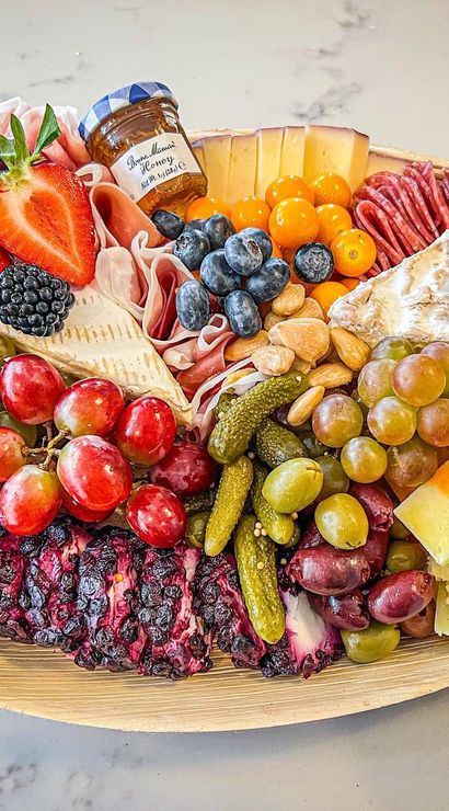 Vibrant gourmet charcuterie board on a wooden platter over a marble countertop with assorted cheeses, rolled salami and prosciutto, strawberries, grapes, blackberries and blueberries, olives, cornichons, nuts and a small jar of honey — colorful party cheese platter.