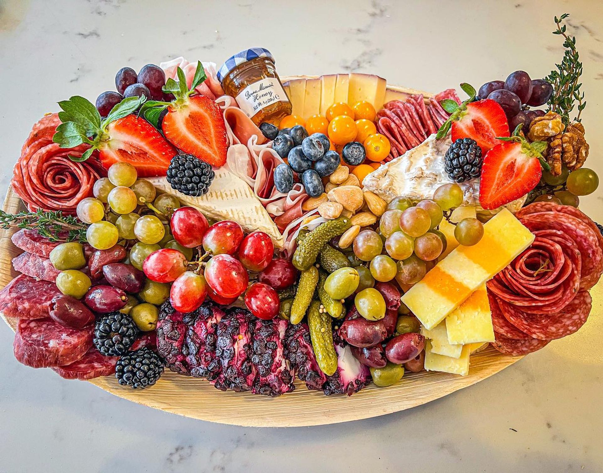 Vibrant charcuterie grazing board on a wooden platter with brie and cheddar, rolled salami roses, grapes, strawberries, blackberries, blueberries, olives, cornichons, nuts and a small jar of jam on a marble countertop