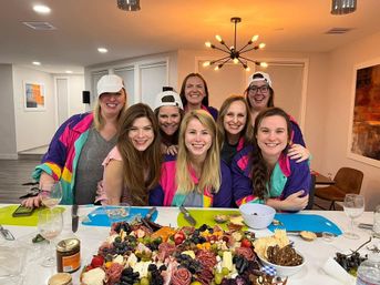 Eight smiling women posing around a large colorful charcuterie board with cheeses, fruits and cured meats in a modern home kitchen and dining area under a starburst pendant light.