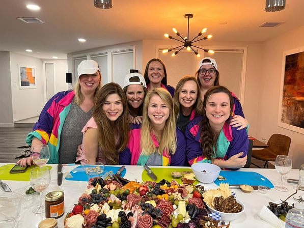 Eight friends smiling around a kitchen island in a modern home, wearing colorful retro windbreakers and caps, with a large charcuterie board of meats, cheeses and fruit.