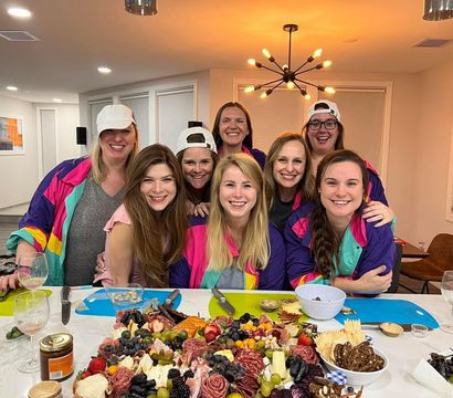 Eight smiling women posing around a large colorful charcuterie board with cheeses, fruits and cured meats in a modern home kitchen and dining area under a starburst pendant light.
