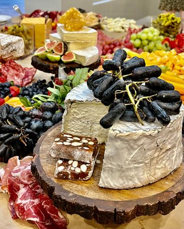 Gourmet charcuterie board on a rustic wooden slab with a wheel of brie topped with long black grapes, fig halves, almond-studded nougat, cured meats, berries and cubed cheeses.