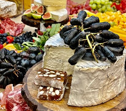 Gourmet charcuterie board on a rustic wooden slab with a wheel of brie topped with long black grapes, fig halves, almond-studded nougat, cured meats, berries and cubed cheeses.