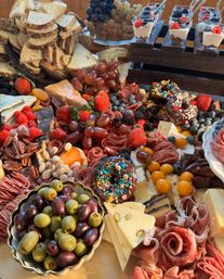 Vibrant charcuterie grazing board with assorted cheeses, rolled cured meats, bowls of green and Kalamata olives, clusters of grapes, strawberries, blackberries, blueberries and raspberries, nuts and dried fruit, sliced multigrain bread, chocolate‑glazed sprinkle donuts, and mini yogurt parfaits.