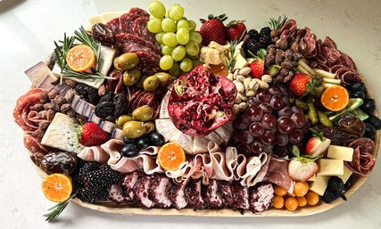 Vibrant charcuterie grazing board bursting with color: green and red grapes, halved pomegranate, strawberries, blackberries, clementines, marinated olives, dates, pistachios, assorted cheeses, rolled cured meats, cornichons and rosemary garnish.