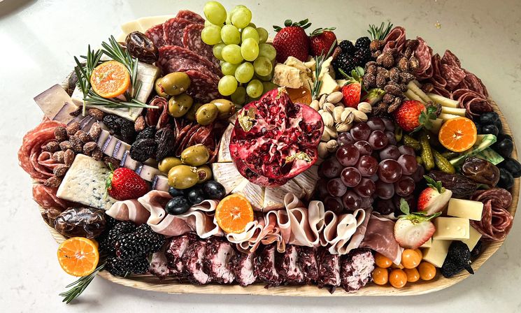 Vibrant gourmet charcuterie board on a wooden platter with a halved pomegranate centerpiece, wheel of brie, blue and marbled cheeses, folded prosciutto and salami rosettes, green and red grapes, olives, pistachios, candied nuts, dates, blackberries, strawberries, mandarin slices and cornichons