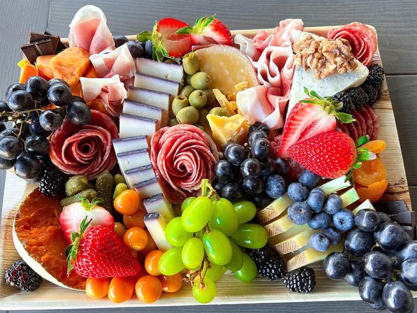Colorful charcuterie board on a wooden platter with assorted cheeses, prosciutto and salami roses, green and black grapes, strawberries, blueberries and blackberries, olives, cornichons, dried apricots, walnuts and dark chocolate.