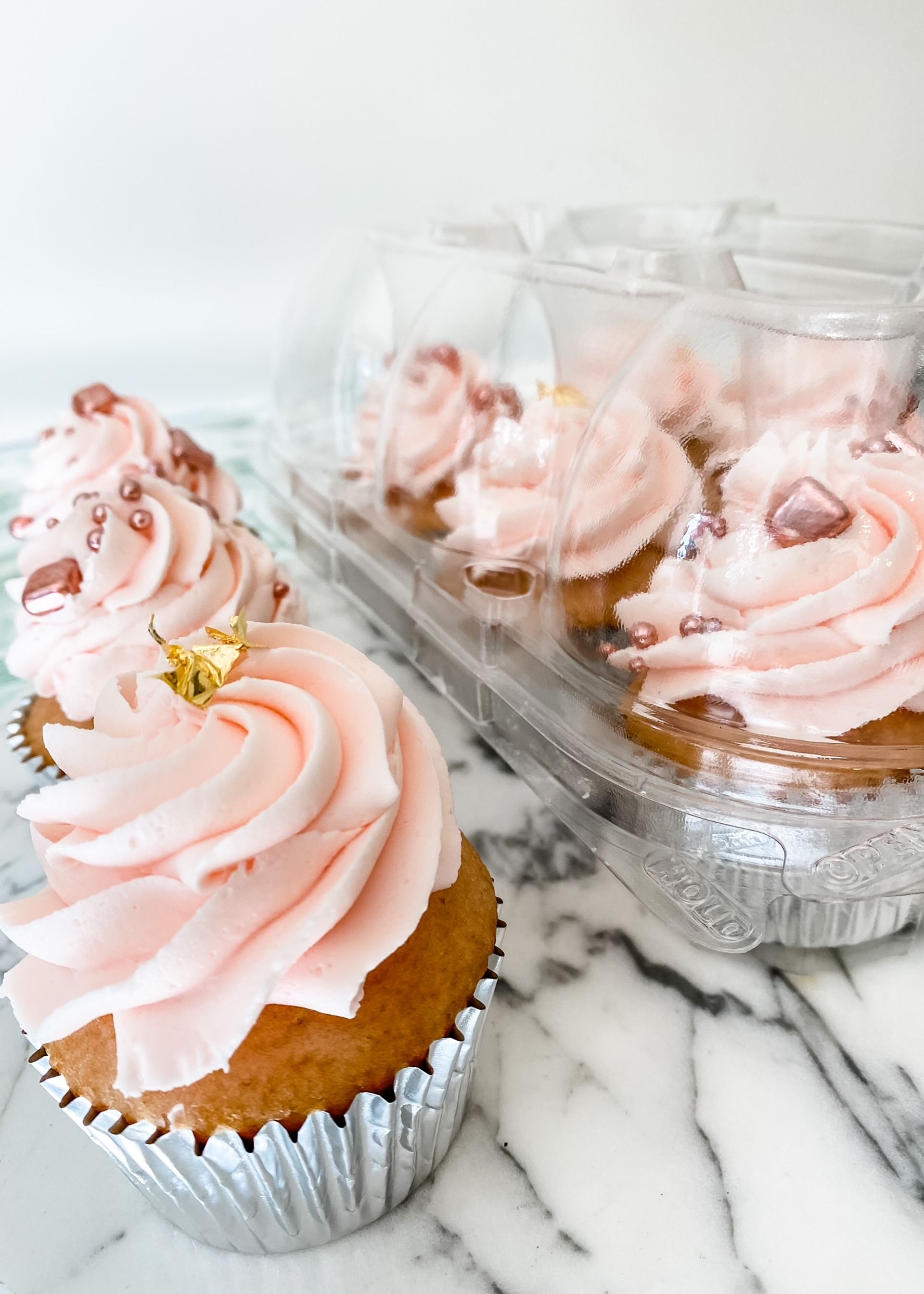 Close-up of pretty pink swirled buttercream cupcakes in silver foil liners, topped with rose-gold sprinkles and edible gold leaf, set on a marble countertop with a clear plastic cupcake container