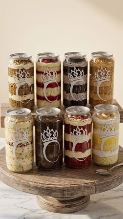 Eight clear can-shaped jars of layered cake desserts (red velvet, chocolate, lemon, funfetti and more) with a decorative crown logo, arranged on a rustic wooden cake stand over a marble countertop.