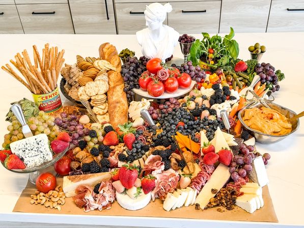 Vibrant overflowing charcuterie grazing board on a modern kitchen island with assorted cheeses, prosciutto and salami, baguette and crackers, breadsticks, olives and pickles, hummus, fresh grapes, strawberries, blueberries, blackberries, cherry tomatoes and a basil centerpiece.