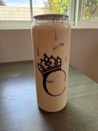 Sunlit window behind a tall reusable tumbler of creamy iced milk tea with ice and foam, black crown logo on the bottle, sitting on a wooden table.