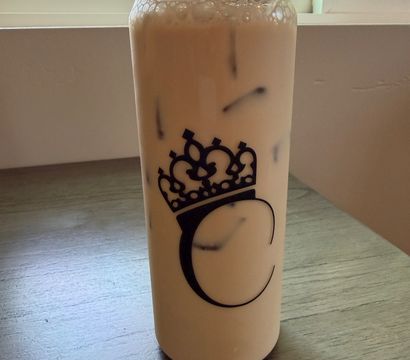 Sunlit window behind a tall reusable tumbler of creamy iced milk tea with ice and foam, black crown logo on the bottle, sitting on a wooden table.