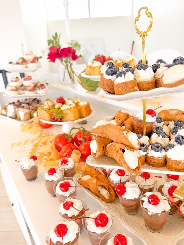Bright dessert table in a sunlit modern setting with a gold tiered stand displaying mini blueberry cream tarts, cannoli filled with cream, chocolate mousse cups topped with whipped cream and cherries, and assorted bite-sized pastries on white platters.