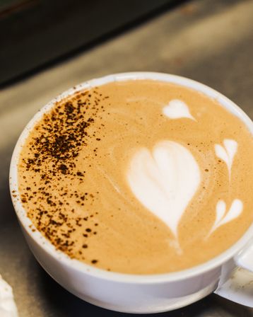 Top-down shot of a creamy latte in a white cup with heart-shaped latte art, two small hearts and cocoa sprinkles along the rim on a café counter.