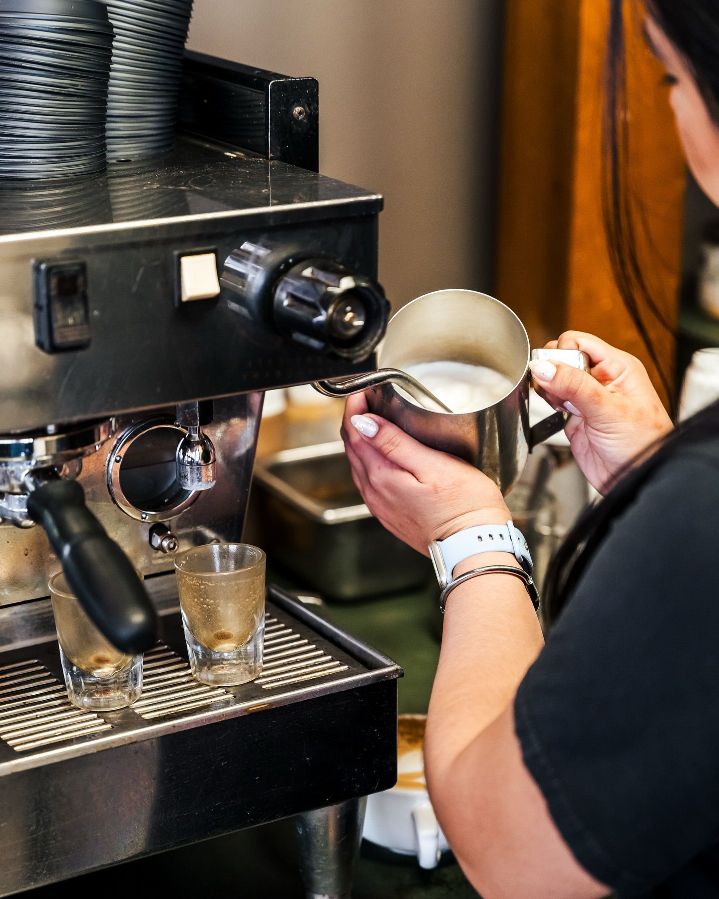 Close-up of a barista steaming milk in a stainless pitcher at a commercial espresso machine, two espresso shot glasses on the drip tray — coffee shop latte-making with steam wand and milk froth.