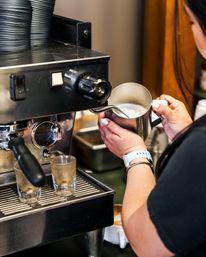 Close-up of a barista steaming milk in a stainless pitcher at a commercial espresso machine, two espresso shot glasses on the drip tray — coffee shop latte-making with steam wand and milk froth.