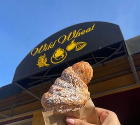 Flaky powdered croissant in a paper sleeve held up in front of a bakery awning with yellow food icons against a bright blue sky