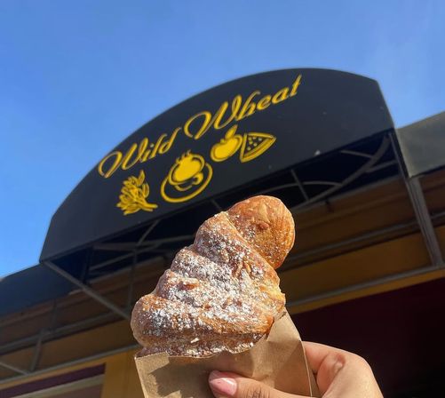 Flaky powdered croissant in a paper sleeve held up in front of a bakery awning with yellow food icons against a bright blue sky