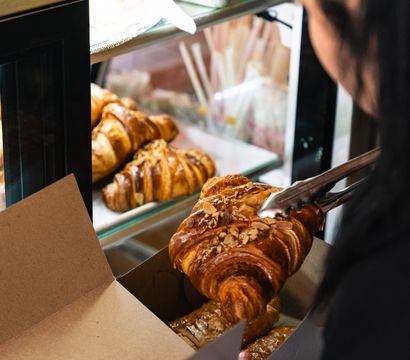 Almond-topped croissant picked up with tongs and placed into a takeaway box at a bakery display case with golden croissants behind it