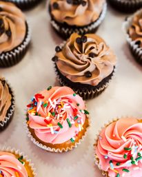 Close-up of assorted cupcakes on a bakery tray — pink swirled frosting topped with colorful sprinkles and chocolate-frosted cupcakes with mini chips
