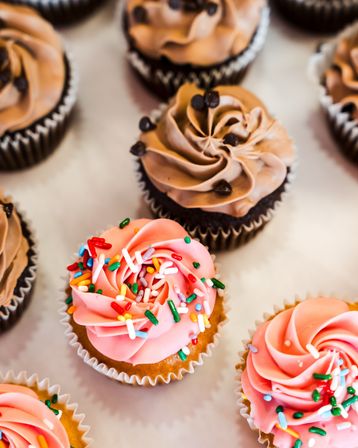 Close-up of assorted cupcakes on a bakery tray — pink swirled frosting topped with colorful sprinkles and chocolate-frosted cupcakes with mini chips