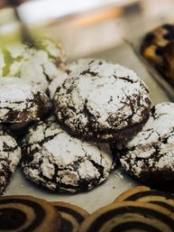 Close-up of chocolate crinkle cookies dusted with powdered sugar on a bakery display tray with spiral pinwheel cookies in the foreground